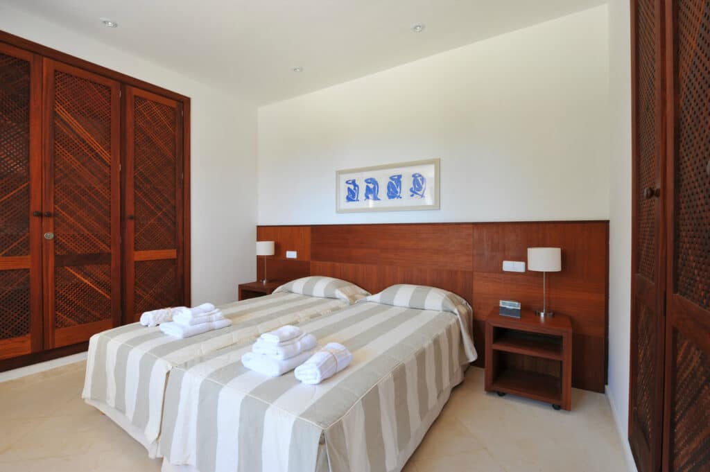 A minimalist bedroom designed for luxury travel, featuring two single beds pushed together and covered in elegant striped linens. Rolled white towels add a touch of refinement. The room boasts wooden paneling, a large wardrobe, and framed artwork above the headboard. David Pearson Travel