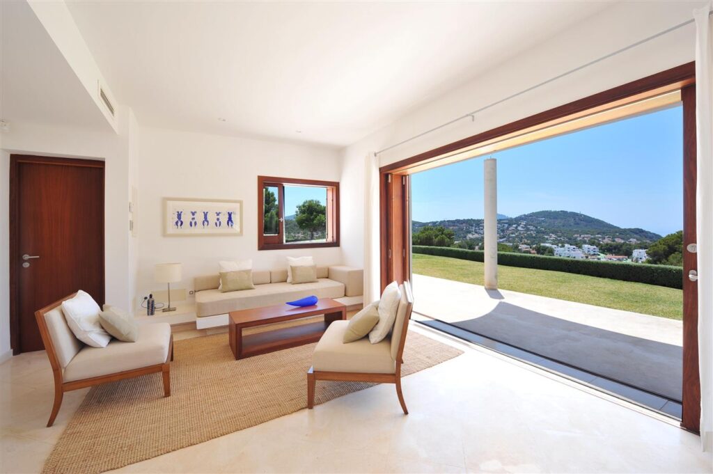 Bright, modern living room with beige furniture and a wooden coffee table on a woven rug. Large sliding glass doors open to a patio, offering views reminiscent of luxury travel destinations with green hills and a clear blue sky. A framed artwork adorns the wall, adding a touch of elegance. David Pearson Travel