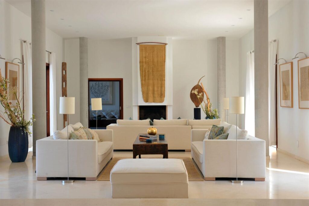A modern living room exudes luxury, with white sofas elegantly arranged around a wooden coffee table. Tall vases with branches accent the sides, while art pieces adorn the walls—a serene space akin to a luxury travel experience, filled with natural light from large windows. David Pearson Travel
