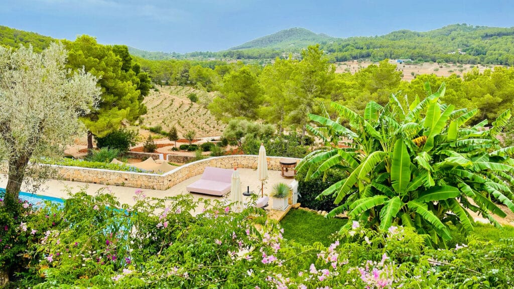 Lush landscape with green hills and a garden featuring banana trees and flowering plants. A stone walkway leads past a patio with an umbrella and seating, epitomizing luxury travel, with a pool partially visible. Hilly terrain stretches into the distance under a cloudy sky. David Pearson Travel