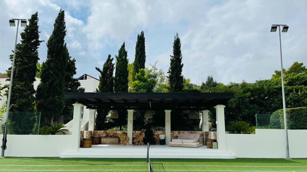 A tennis court with a white and green playing surface graces the foreground, embodying the essence of luxury travel. Behind it, a covered outdoor seating area with a couch and table invites relaxation amidst tall trees and lush greenery beneath a cloudy sky. David Pearson Travel