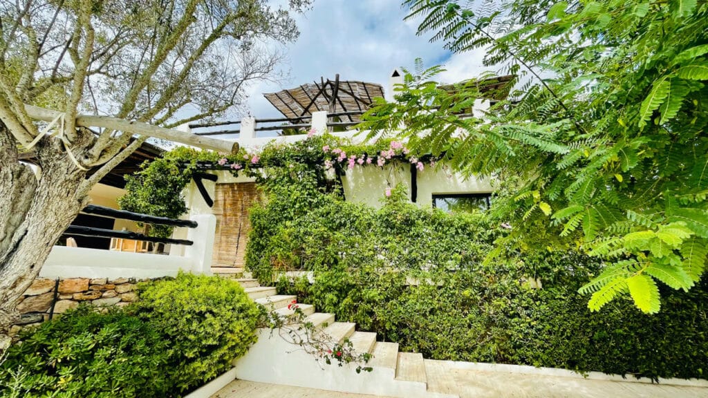This Mediterranean-style home, perfect for luxury travel enthusiasts, is nestled in lush greenery. It features a white exterior, wooden accents, and a terrace. Stone steps lead to the entrance adorned with flowering plants and framed by trees under a partly cloudy sky. David Pearson Travel