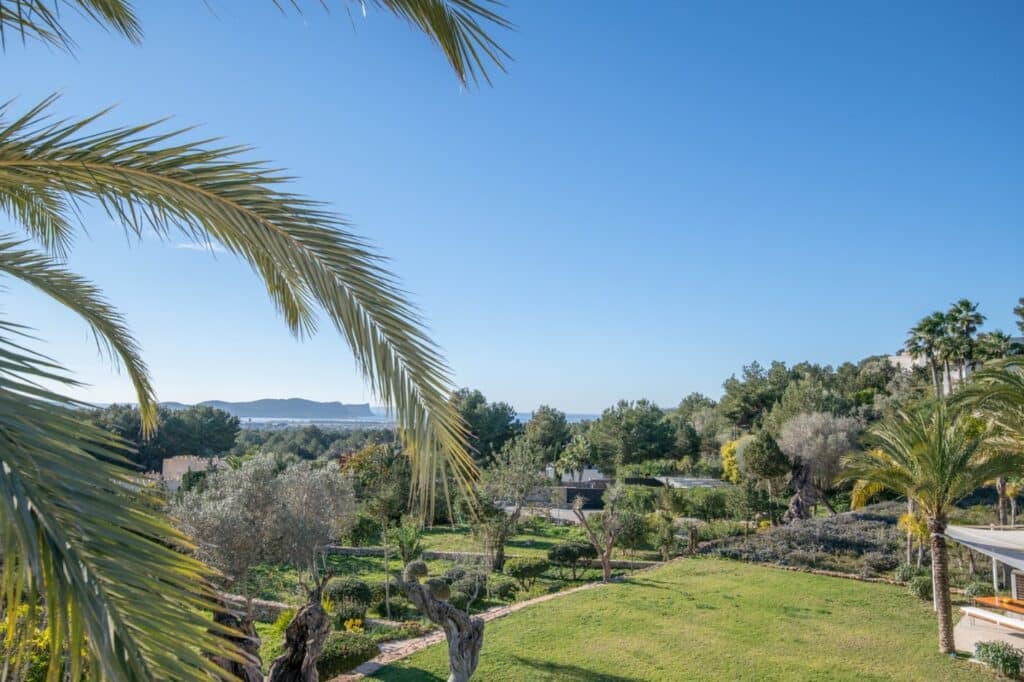 A scenic view of a lush green garden with palm trees and neatly trimmed grass. Olive trees and other greenery extend toward a distant hill, under a bright blue sky. A glimpse of the sea is visible on the horizon, offering a touch of luxury travel to this idyllic setting. David Pearson Travel