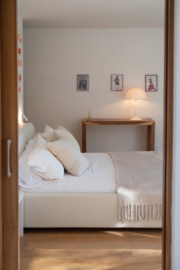 A cozy bedroom exudes the charm of luxury travel, with a white bed piled high with pillows and a beige throw. A wooden side table with a lamp sits to the right, while three small pictures hang above. The warm lighting and wooden flooring enhance this serene escape. David Pearson Travel