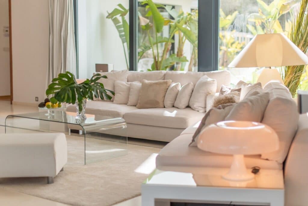 The bright, luxurious living room features a large white sectional sofa adorned with neutral cushions. A glass coffee table with a plant in a vase complements the space. Floor-to-ceiling windows reveal lush greenery outside, creating an atmosphere reminiscent of luxury travel. David Pearson Travel