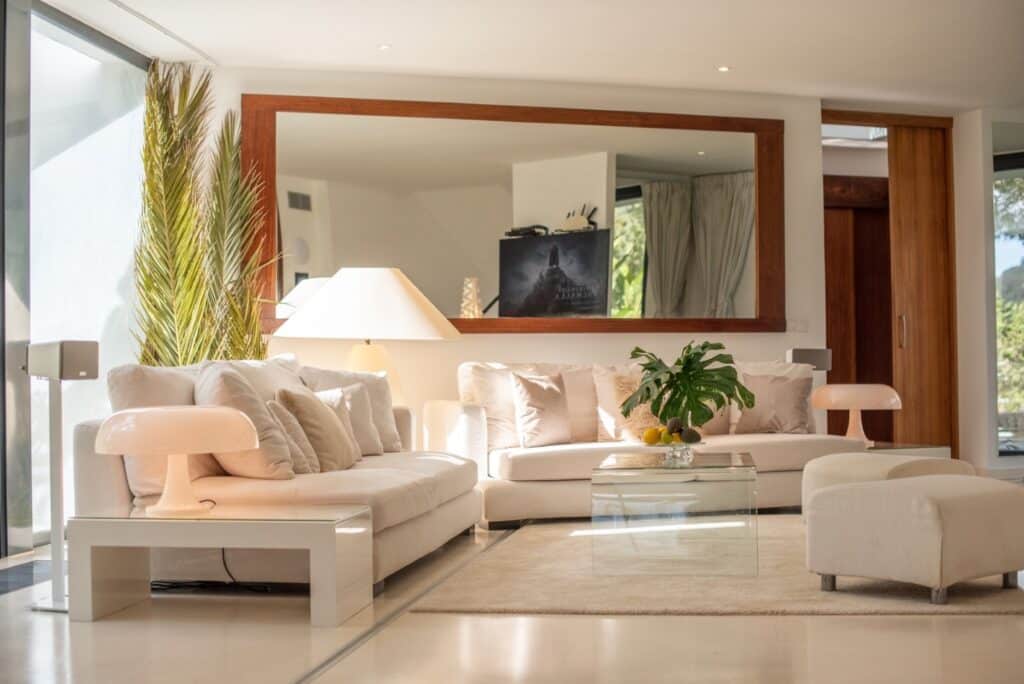 A modern living room evokes the essence of luxury travel with white sofas, a glass coffee table on a light rug, and a mirror reflecting natural light. Touches of green plants and a bowl of lemons add color, while a floor lamp and abstract artwork complete the sophisticated décor. David Pearson Travel
