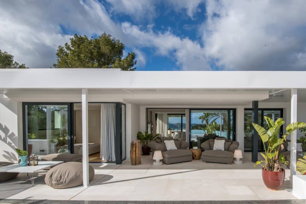 Experience luxury travel in this modern open-plan living space with glass sliding doors leading to a terrace featuring outdoor seating. Natural light floods in, highlighting the minimalist décor, while a pool and palm trees bask under a partly cloudy sky. David Pearson Travel