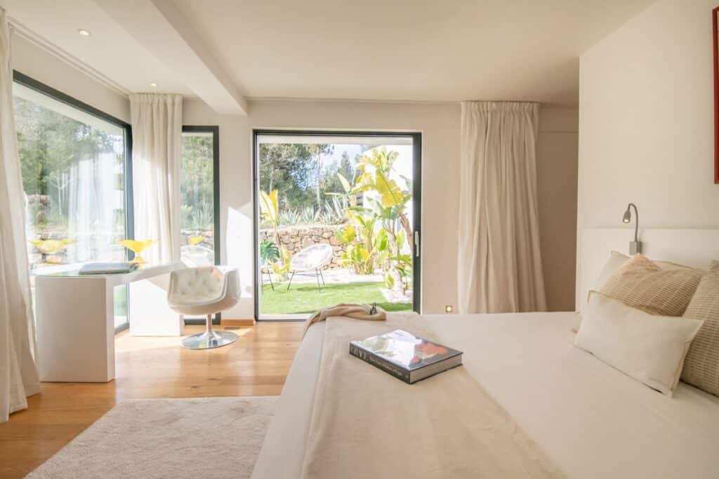 Bright, modern bedroom with white decor, featuring a large bed and a small desk and chair. Two large windows open to a garden with leafy plants, inviting natural light to fill this luxury travel-inspired space. A book rests on the bed. David Pearson Travel