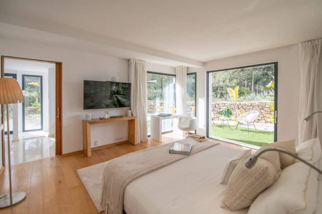 Spacious bedroom with large windows, a double bed, a wall-mounted TV, and a wooden desk. Light wooden flooring complements the neutral decor, ideal for luxury travel seekers. Outside, a lush garden and stone wall are visible. A warm, cozy atmosphere fills the room. David Pearson Travel