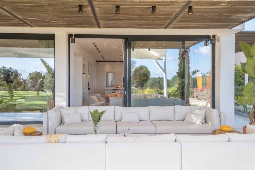 Luxurious and inviting, this spacious patio boasts a large white sectional sofa facing glass sliding doors. Behind the doors, an open-plan living area is visible. Greenery envelops the scene, while a wooden ceiling adds rustic charm. Perfect for those seeking a sophisticated luxury travel experience at home. David Pearson Travel