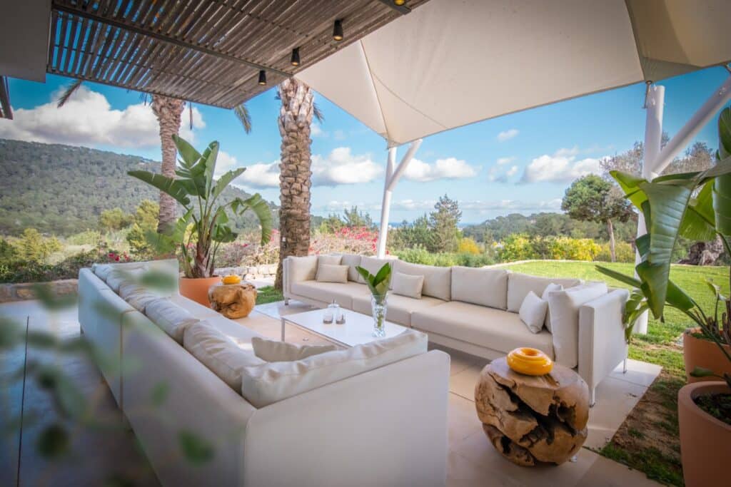 The outdoor seating area features plush white sofas under a canopy, perfect for luxury travel relaxation. Surrounded by lush greenery and palm trees, a wooden coffee table holds tasteful decorative items. The view showcases majestic mountains and a blue sky with scattered clouds. David Pearson Travel