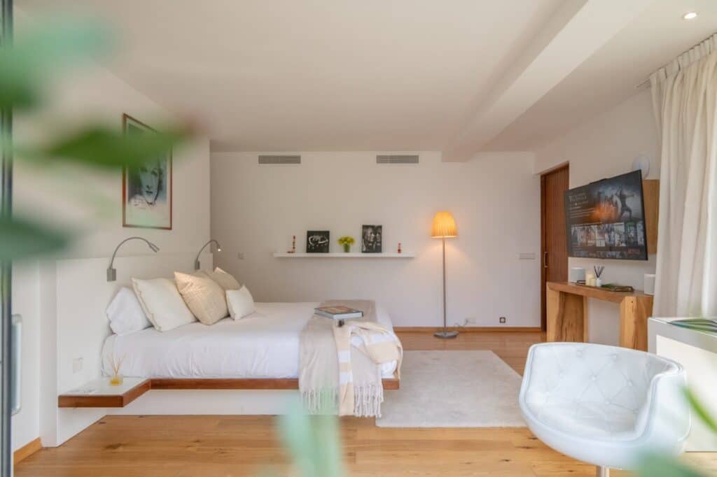 A spacious, modern bedroom ideal for luxury travel, boasting wooden floors and a large bed with white linens and a beige throw. A TV on a wooden stand, cozy armchair, and shelves with decorations enhance the space, warmly lit by a standing lamp. David Pearson Travel