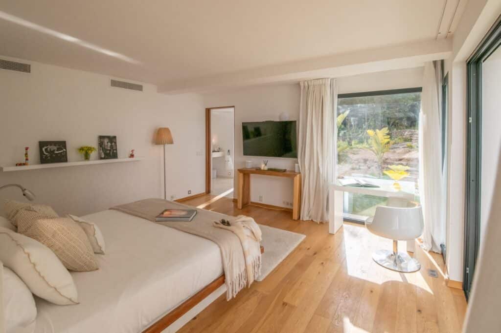 A bright bedroom with a large window offering a scenic view, perfect for luxury travel experiences. It features a neatly made bed with cushions, a wooden desk with a lamp and TV, a modern chair, and decorative items on a shelf. Light wood flooring adds warmth to the room. David Pearson Travel