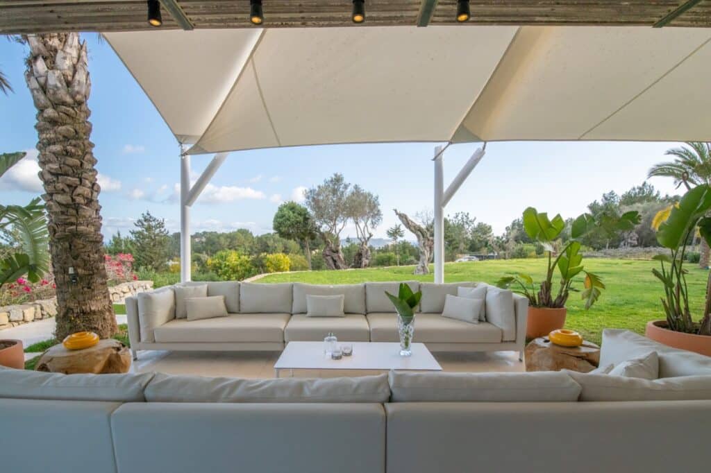 The outdoor patio setup offers a luxury travel vibe with beige sectional sofas under a white canopy, overlooking a lush green garden. Accent tables adorned with decorative vases and plants enhance the sunny, serene scene, featuring palm trees and vibrant flowering plants. David Pearson Travel