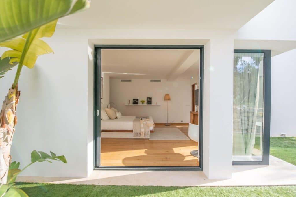 A bright and modern bedroom, reminiscent of luxury travel suites, is viewed through large sliding glass doors. The room features a bed with neutral bedding, a wooden chair, and a standing lamp. A potted plant outside on the grass complements the minimalist design. David Pearson Travel