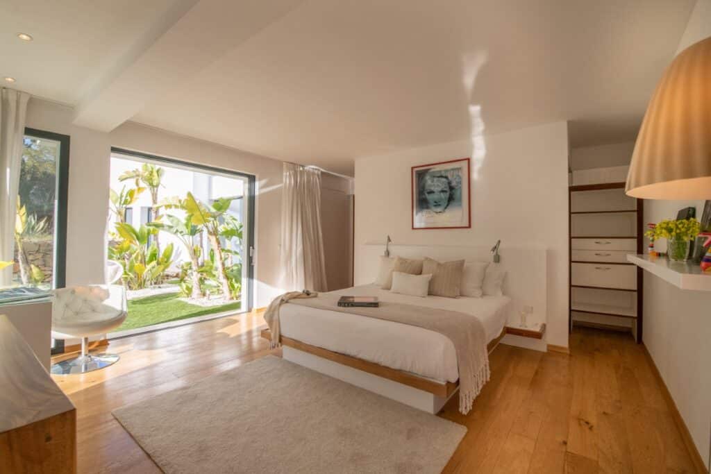 Spacious bedroom with wooden floors, featuring a large bed with beige and white bedding reminiscent of luxury travel accommodations. Theres a portrait above the bed, a chair by a window with a garden view, and a large pendant light overhead. Open shelves visible on the right. David Pearson Travel