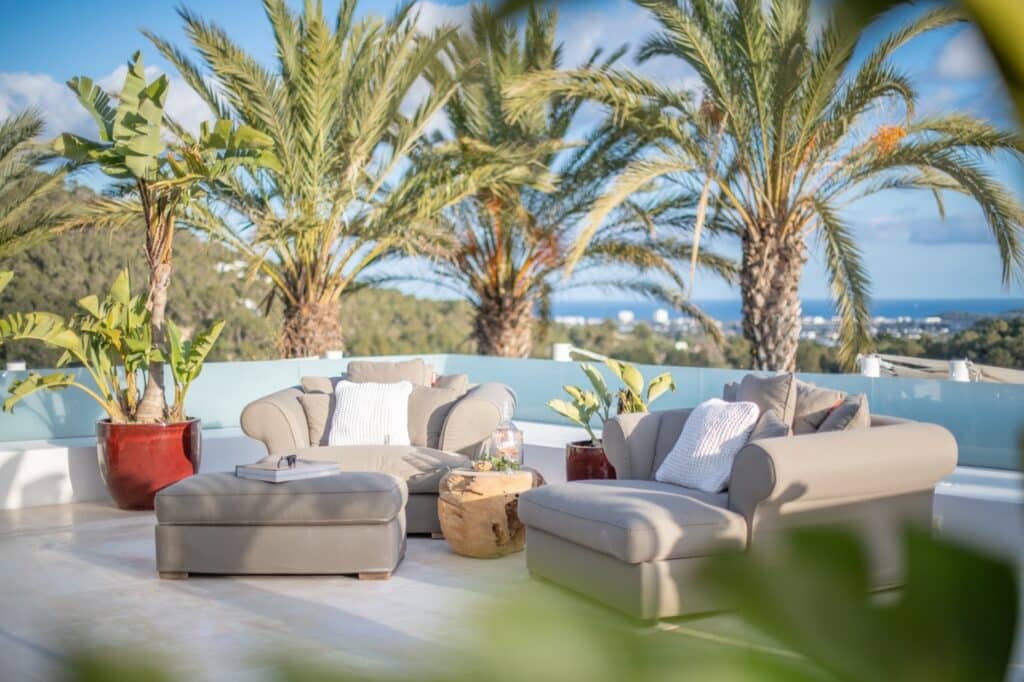 A rooftop terrace with beige cushioned chairs and a matching ottoman, perfect for luxury travel relaxation, surrounded by large palm trees in red pots. A round wooden stump serves as a coffee table. The sky is blue, offering a distant view of the ocean. David Pearson Travel