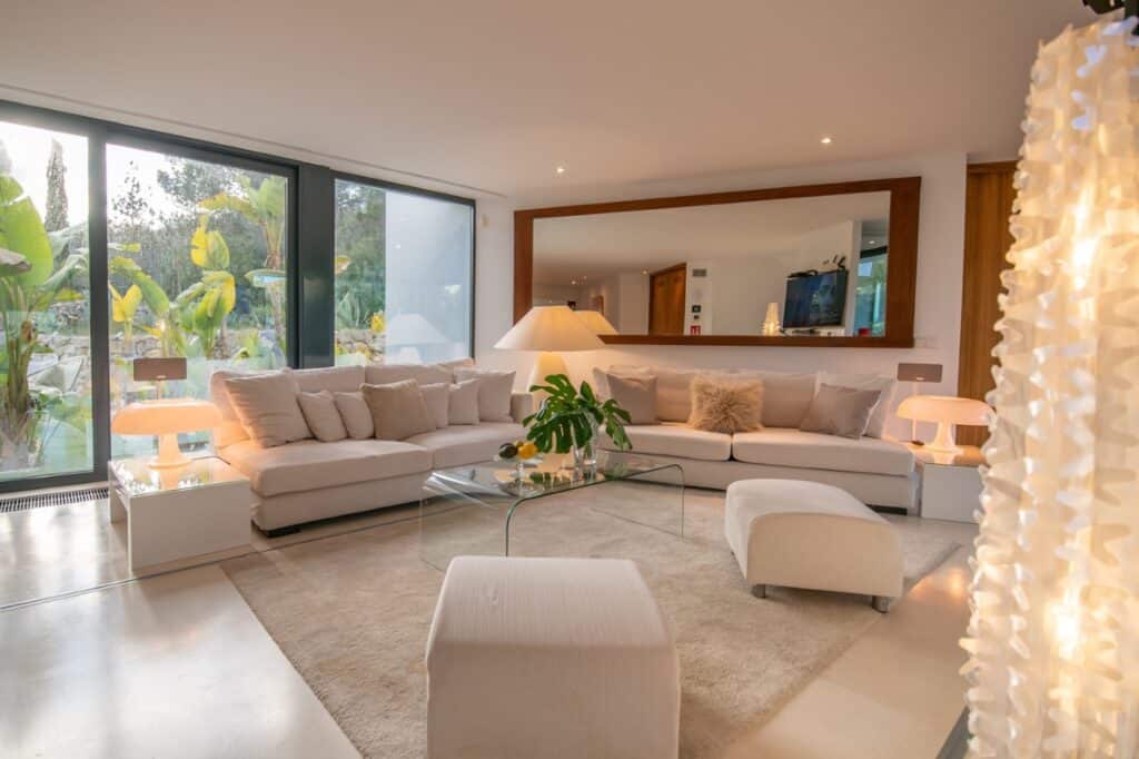 A haven of modern elegance, this living room boasts a large white sectional sofa, complemented by a glass coffee table and plush ottomans. A big mirror and vibrant plants enrich the decor. Floor-to-ceiling windows offer views of lush greenery, creating an ambiance that echoes luxury travel. David Pearson Travel