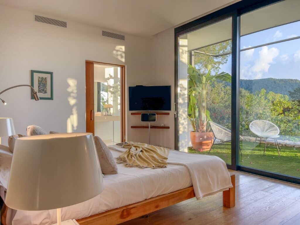A sunlit bedroom with a wooden bed, white bedding, and two lamps exudes a luxury travel vibe. Large floor-to-ceiling windows unveil lush greenery and hills. A TV is mounted on the wall, with a doorway to a modern bathroom. The ambiance is serene and sophisticated. David Pearson Travel