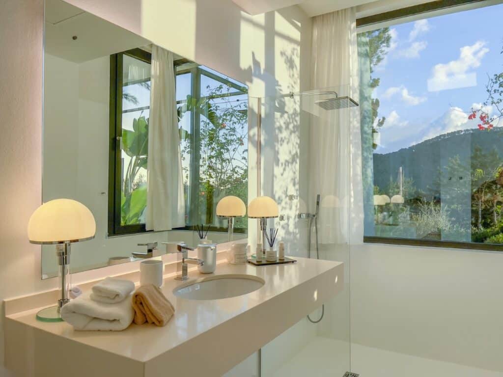 Modern bathroom with a large window displaying scenic mountain and tree views, reminiscent of luxury travel destinations. The room boasts a sleek white countertop with a sink, decorative lamps, folded towels, and a glass-walled shower area. Sunlight streams in, creating a serene ambiance. David Pearson Travel