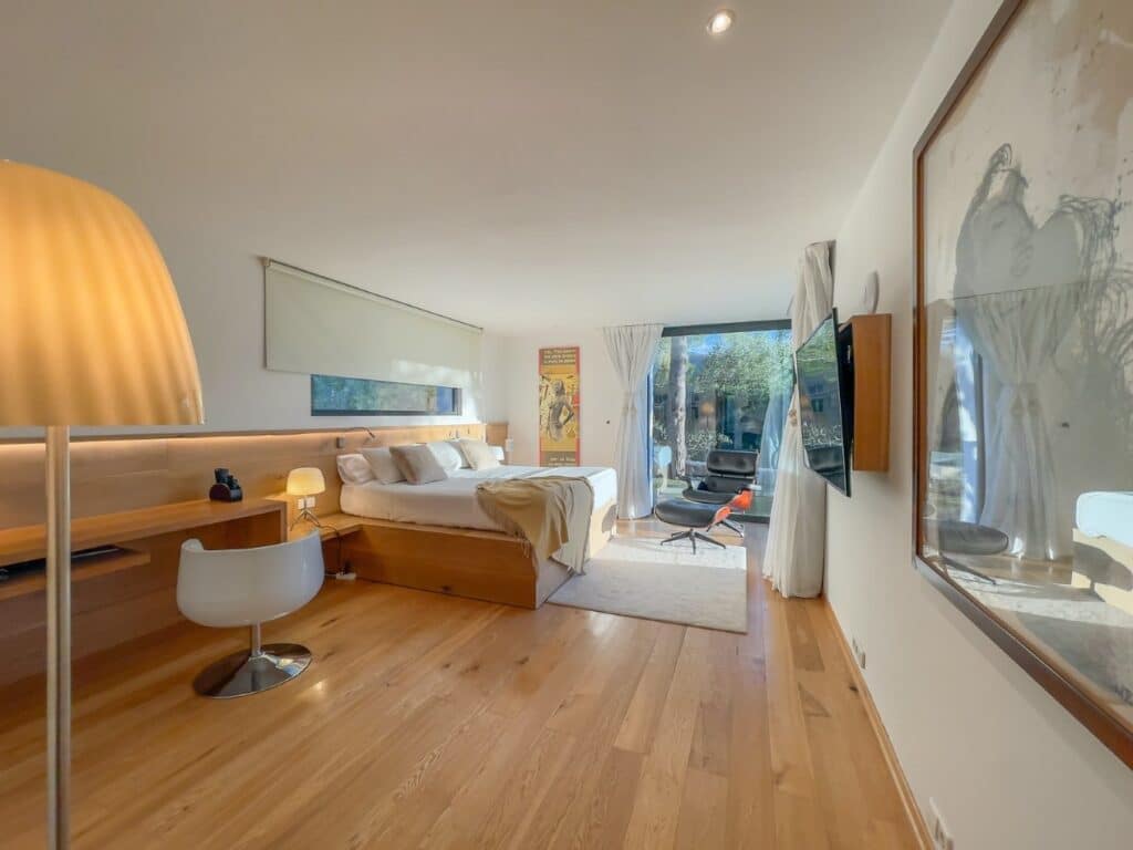A modern, sunlit bedroom epitomizes luxury travel with its large bed, crisp white linens, and elegant wooden headboard. A desk with a white chair sits on the left, while a TV is mounted on the wall. Glass doors open to a serene garden. Warm wooden flooring and contemporary decor complete this tranquil retreat. David Pearson Travel