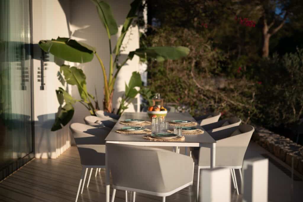 A sunlit outdoor dining area exudes luxury travel vibes with a sleek rectangular white table set for six. Plates and glassware shine under the sunlight, surrounded by elegant gray chairs. Lush banana plants add greenery, while a central glass vase brimming with fruit completes this serene retreat. David Pearson Travel
