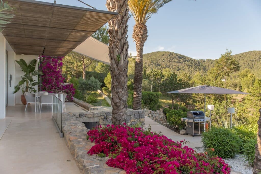 The stylish terrace of this modern house offers a luxury travel experience, with a dining area draped in vibrant bougainvillea. Palm trees and lush greenery envelop the space, while a barbecue grill under an umbrella provides panoramic views of the scenic hills against a clear sky. David Pearson Travel
