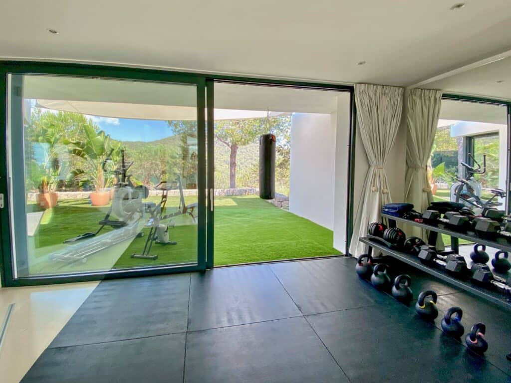 The modern home gym, inspired by luxury travel, features kettlebells and weights neatly arranged on the right. A glass door opens to a patio with artificial grass, plants, outdoor exercise equipment, and a punching bag. Bright natural light floods this exquisite exercise haven. David Pearson Travel