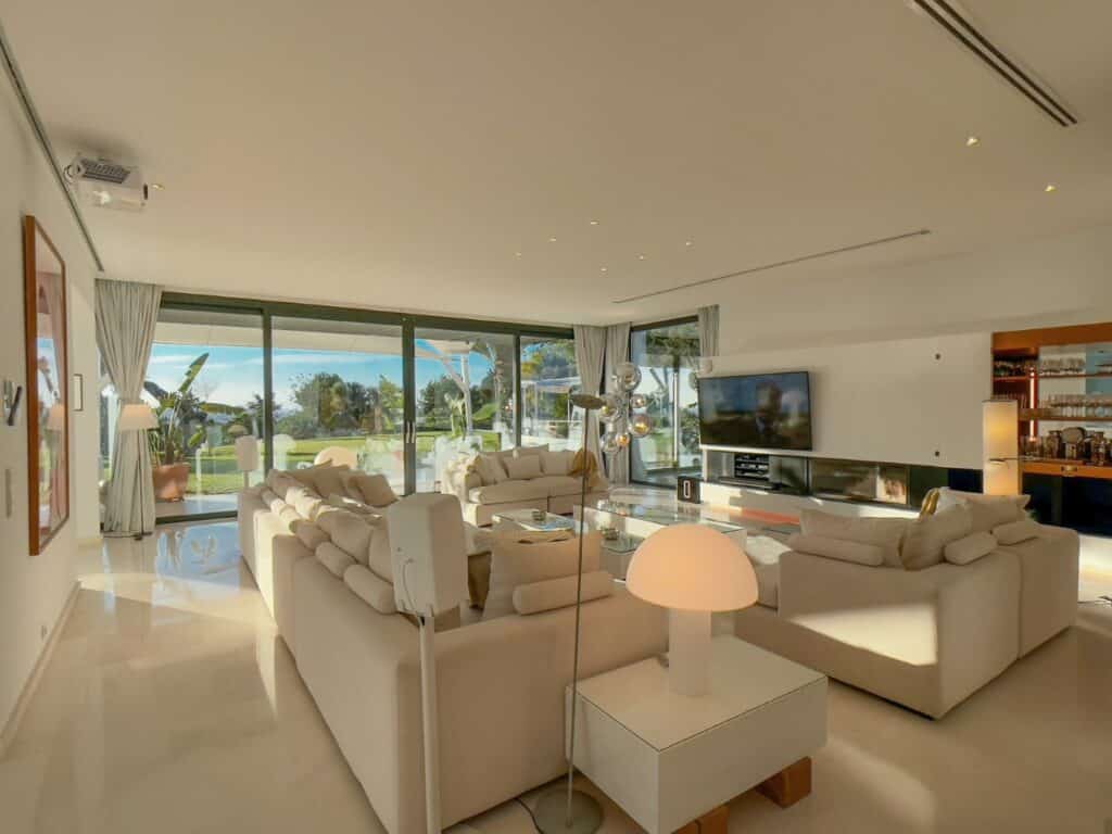 Spacious living room with light-colored sofas and a glass coffee table. Floor-to-ceiling windows offer a view of a garden with palm trees, reminiscent of luxury travel destinations. The room features a wall-mounted TV, modern decor, and ample natural light. David Pearson Travel