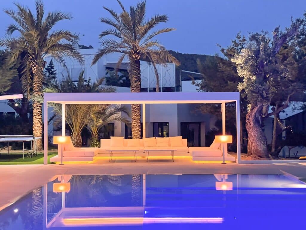 Experience luxury travel at its finest with a sumptuous outdoor seating area by a shimmering pool at dusk. Under a white pergola adorned with glowing lamps, cushioned couches invite relaxation. Towering palm trees and sleek modern architecture complete this idyllic evening escape. David Pearson Travel