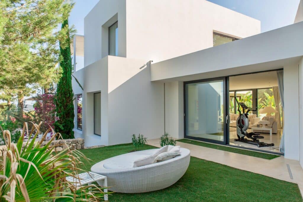 Modern white house with large windows and sliding glass doors leading to a patio. Perfect for luxury travel seekers, the yard features a wicker lounge chair on a grassy area surrounded by plants and trees. Inside, theres a stationary bike visible through the open doors. David Pearson Travel