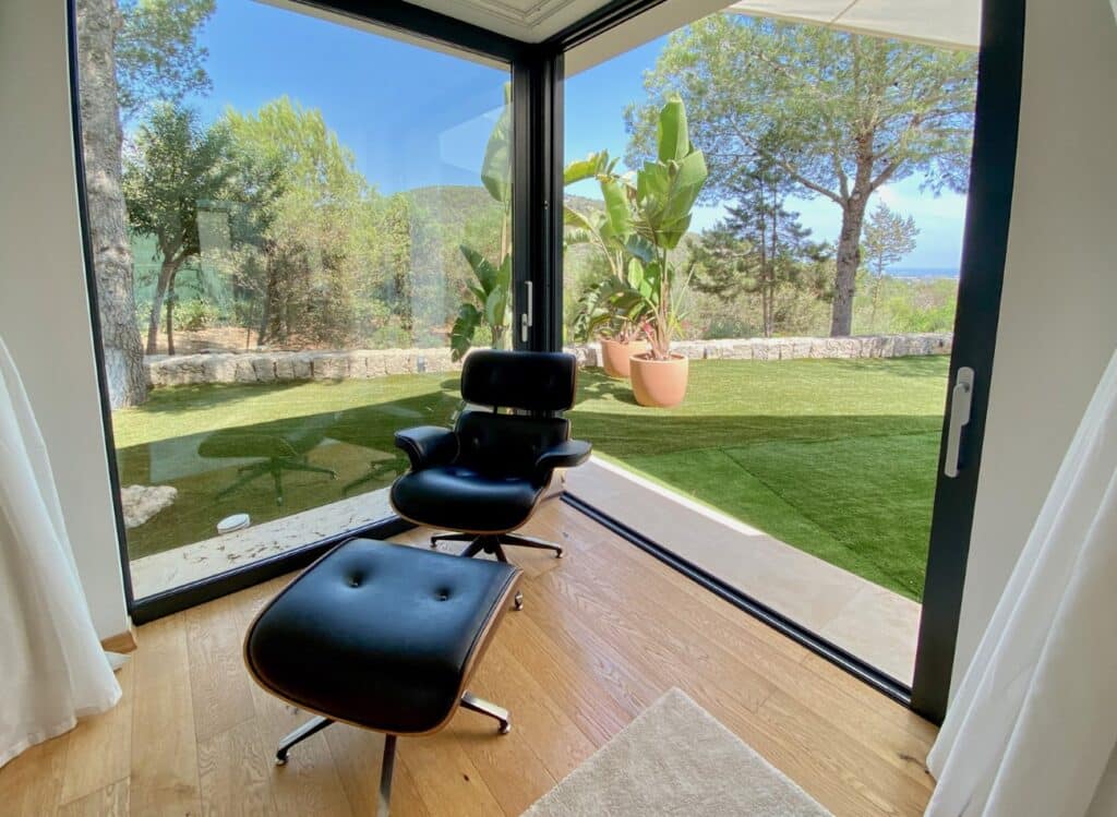 A modern black lounge chair with an ottoman sits in a bright room with large glass windows, evoking the feel of luxury travel. The view outside reveals a lush garden framed by trees and large potted plants, set against a clear blue sky. The wooden floor adds warmth to the elegant design. David Pearson Travel