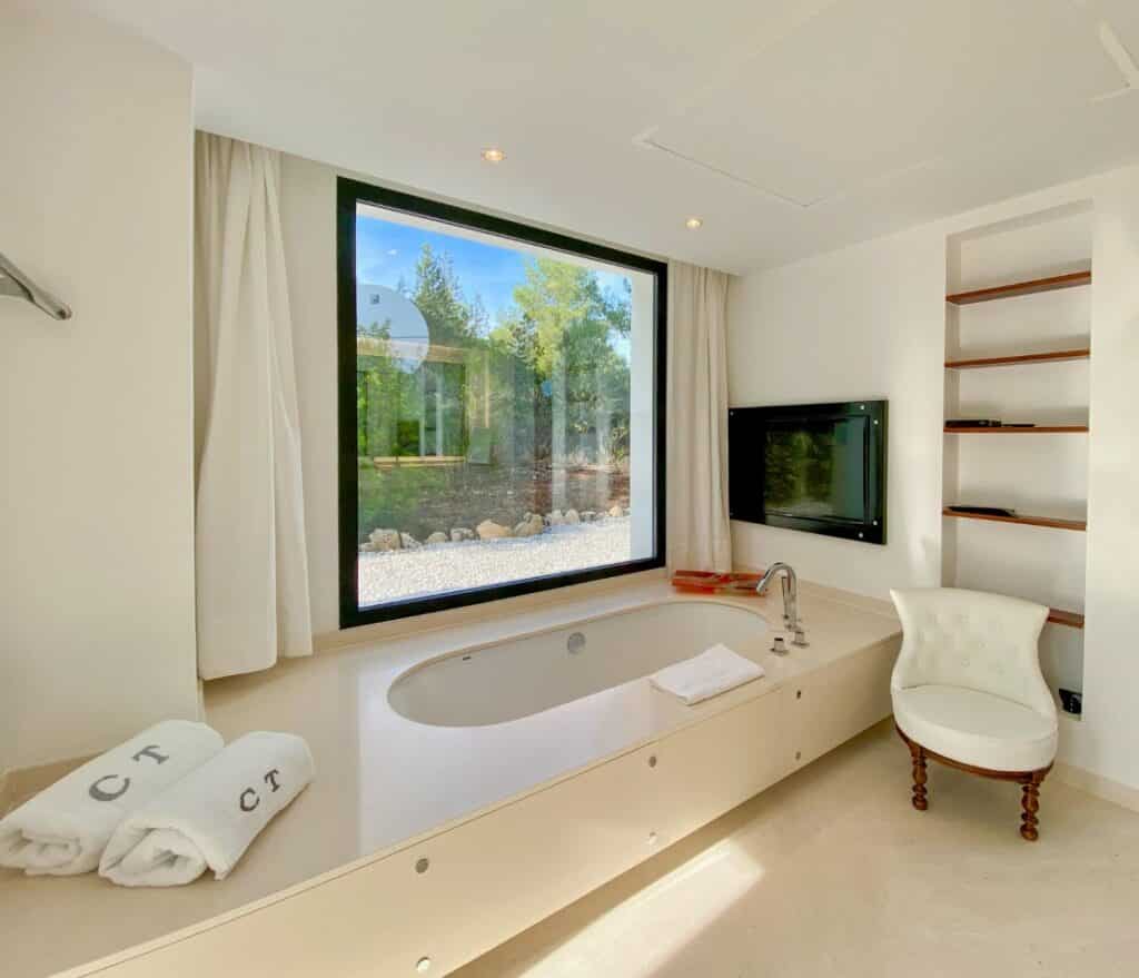 Experience the essence of luxury travel in this modern bathroom, where a large window frames lush greenery. Indulge in a sunken bathtub surrounded by white towels embroidered with CT, enjoy shows on the flat-screen TV, unwind on a cushioned chair, and admire the elegant wooden shelves bathed in natural light. David Pearson Travel