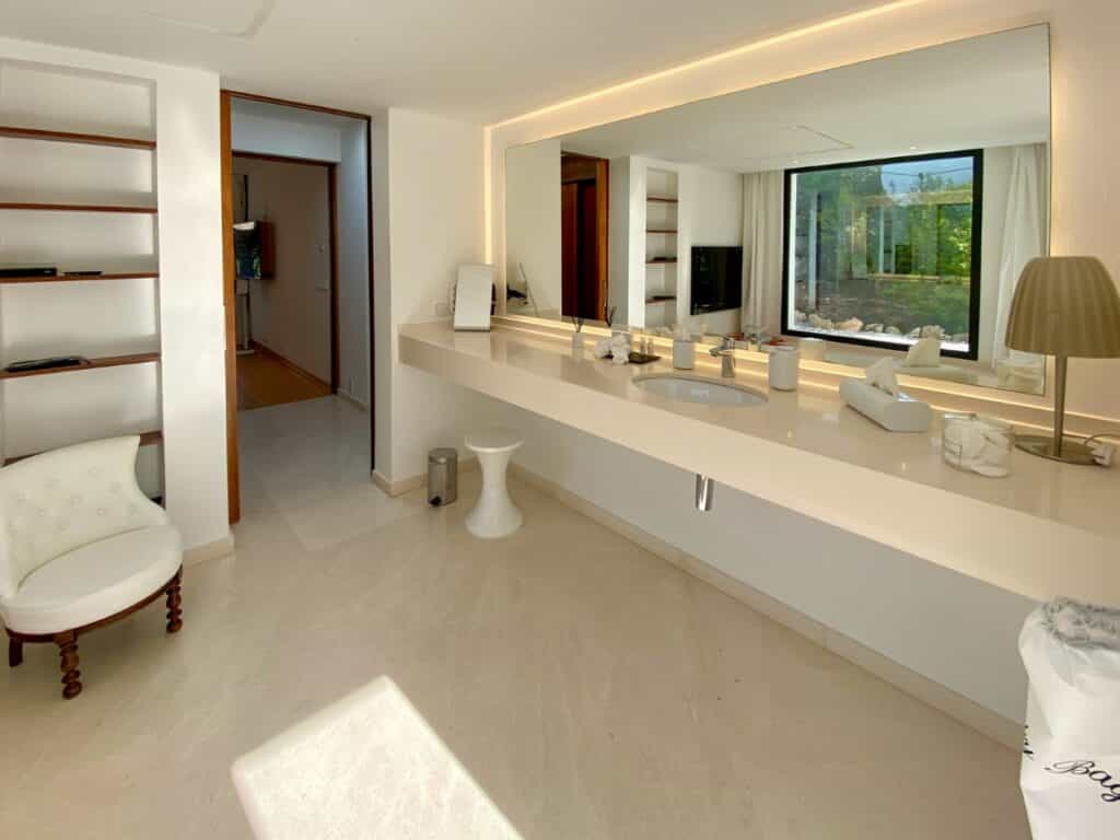 Spacious modern bathroom featuring a large mirror and long countertop with a sink. The room, echoing the elegance of luxury travel, has white walls, shelves, and a white chair with wooden legs. Natural light enters through a large window, highlighting the minimalist decor. David Pearson Travel