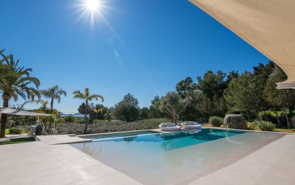 Experience luxury travel at its finest with a modern outdoor pool, surrounded by palm trees and greenery under a clear blue sky. The sun casts shimmering reflections on the water, while comfortable seating offers relaxation in this exquisite setting. David Pearson Travel