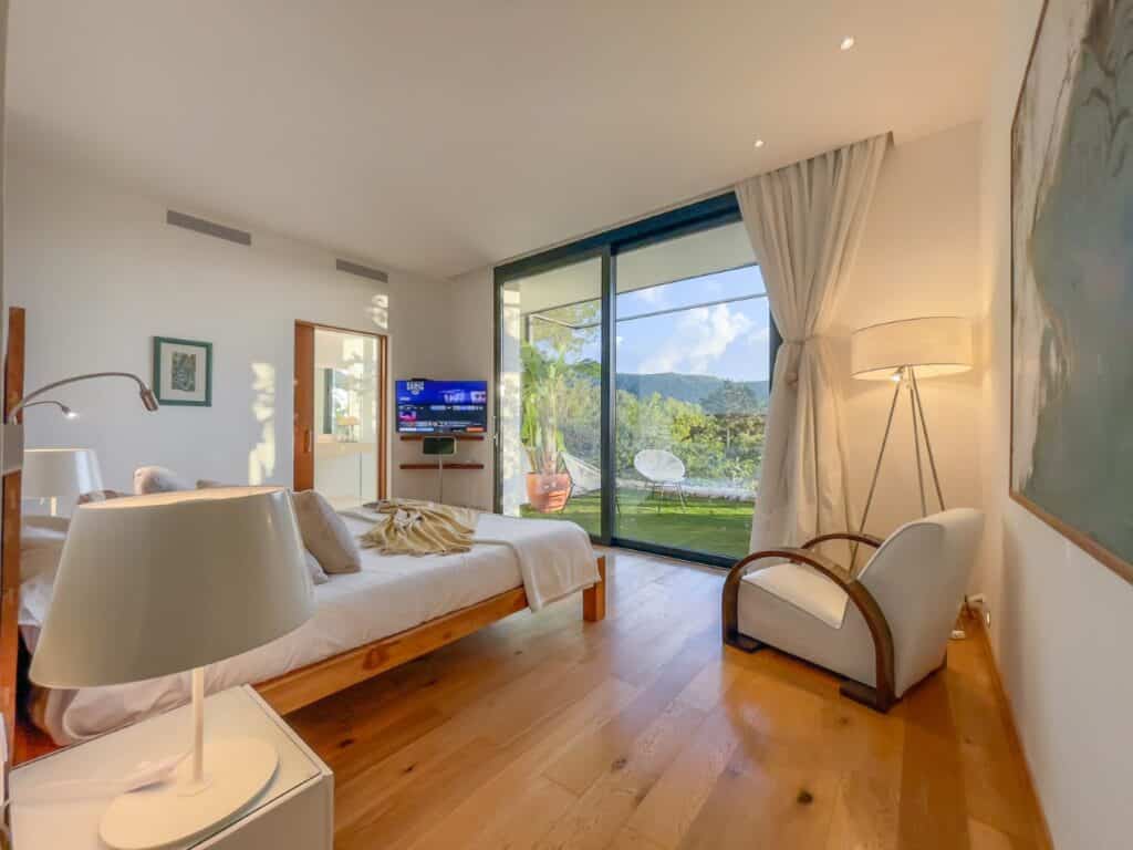 A cozy bedroom with a wooden floor and a large bed facing a flat-screen TV offers the perfect retreat for luxury travel. A floor lamp stands beside an armchair, complemented by a large window revealing a scenic view of mountains and greenery. White curtains are partially drawn. David Pearson Travel