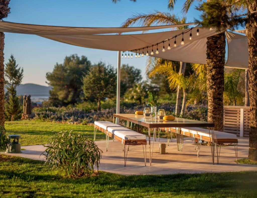 A sunlit outdoor patio epitomizes luxury travel, featuring a long dining table set with glasses and lemons, surrounded by benches. Overhead, string lights are draped across sail-like shades. The area is flanked by palm trees and overlooks a lush, scenic landscape. David Pearson Travel