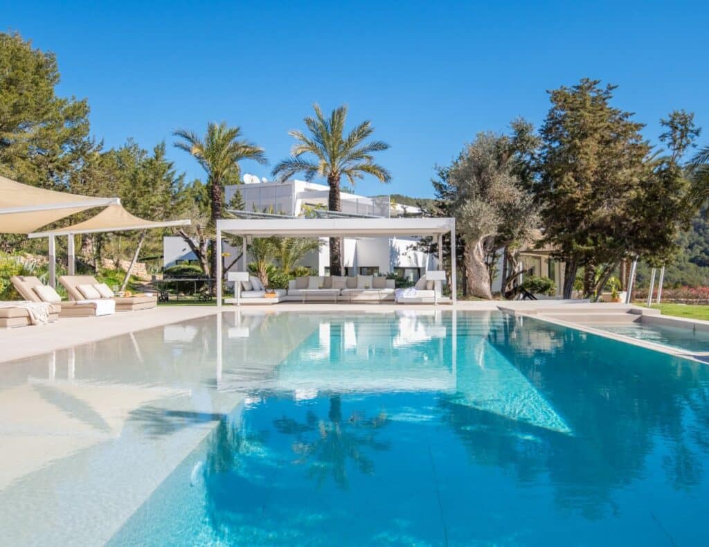 Experience the epitome of luxury travel with a stunning outdoor pool surrounded by palm trees and sun loungers under shade canopies. A sleek, modern villa graces the background, perfectly blending elegance and relaxation against a clear blue sky. David Pearson Travel