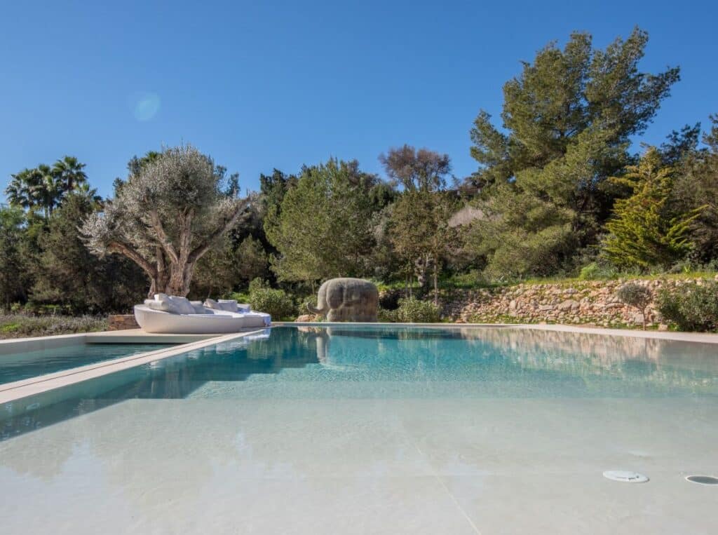 A serene outdoor swimming pool with clear blue water is surrounded by lush trees and greenery, epitomizing luxury travel. A lounge area featuring white cushioned seating is elegantly placed on a platform extending over the water. The sky is clear and blue. David Pearson Travel