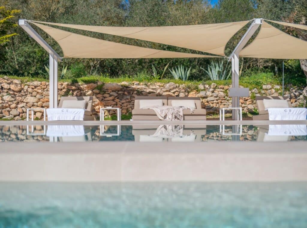 A sunny, luxurious poolside scene with reflective water, lounge chairs, and beige fabric canopies. The background features a stone wall, lush greenery, and a clear blue sky, epitomizing the essence of luxury travel in this tranquil outdoor setting. David Pearson Travel
