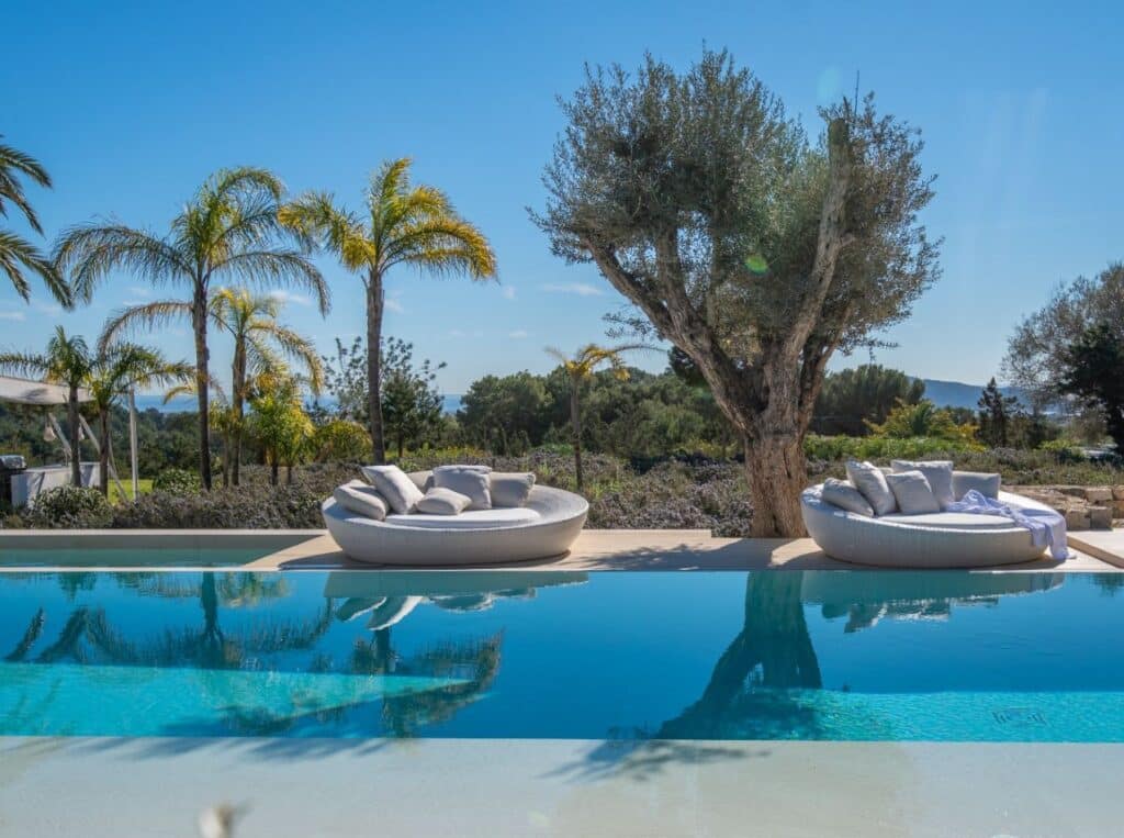 Luxury Travel experience: Outdoor pool scene with two round, cushioned lounge chairs beside a clear blue pool. Olive tree and palm trees in the background under a bright blue sky. A scenic landscape offers greenery in the distance. David Pearson Travel