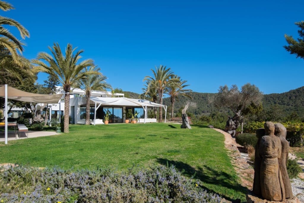 Experience luxury travel at its finest with this modern white house, featuring large windows and surrounded by palm trees and lush greenery. A well-maintained lawn stretches to a stone path, while wooden sculptures grace the foreground against a backdrop of blue sky and wooded hills. David Pearson Travel