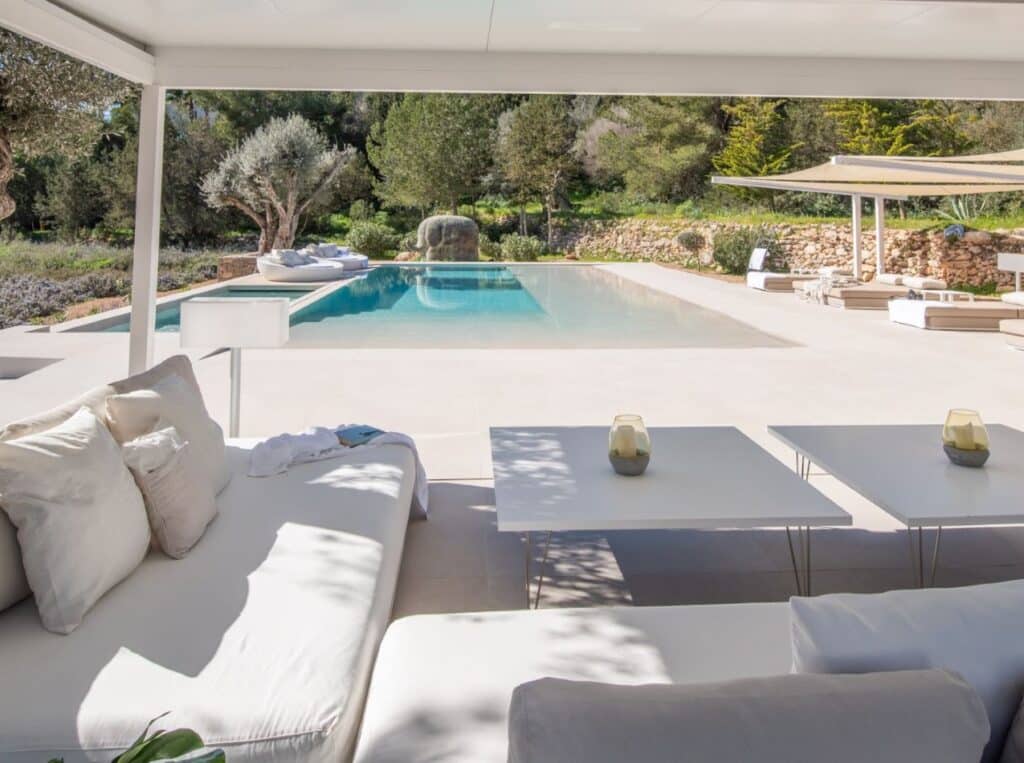 A luxurious outdoor patio, perfect for a luxury travel experience, features white cushioned seating overlooking a large rectangular pool. The tranquil scene is embraced by greenery and trees, with sunlight casting gentle shadows. Candles rest on white tables by the seating area. David Pearson Travel