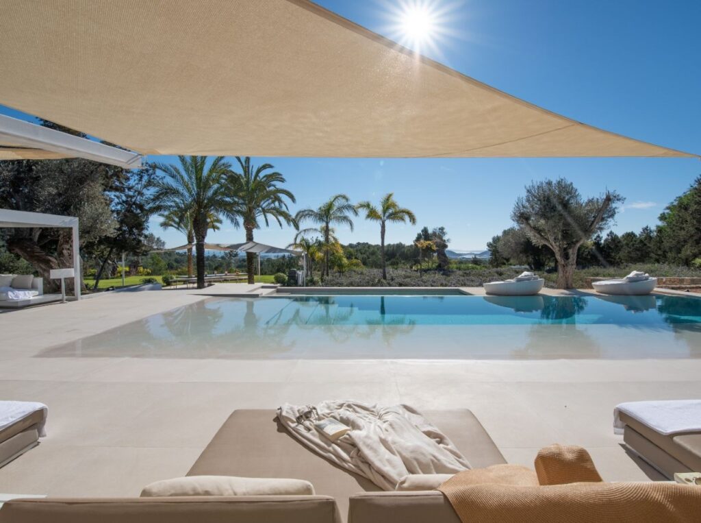 Experience luxury travel at its finest in this outdoor oasis, featuring beige lounge chairs and a chic sunhat. The pool mirrors the clear blue sky and palm trees, while a sunshade casts elegant shadows across the scene, complemented by distant hills and lush greenery. David Pearson Travel