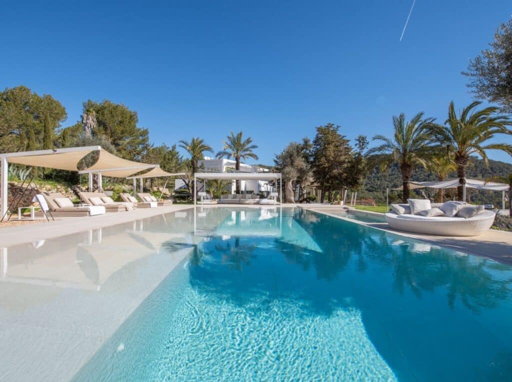Indulge in luxury travel at this exquisite outdoor pool area, where clear blue waters meet the elegance of palm trees and canopy-covered lounge chairs. A modern white villa graces the background, framed by lush greenery and a clear sky with a striking jet trail. David Pearson Travel