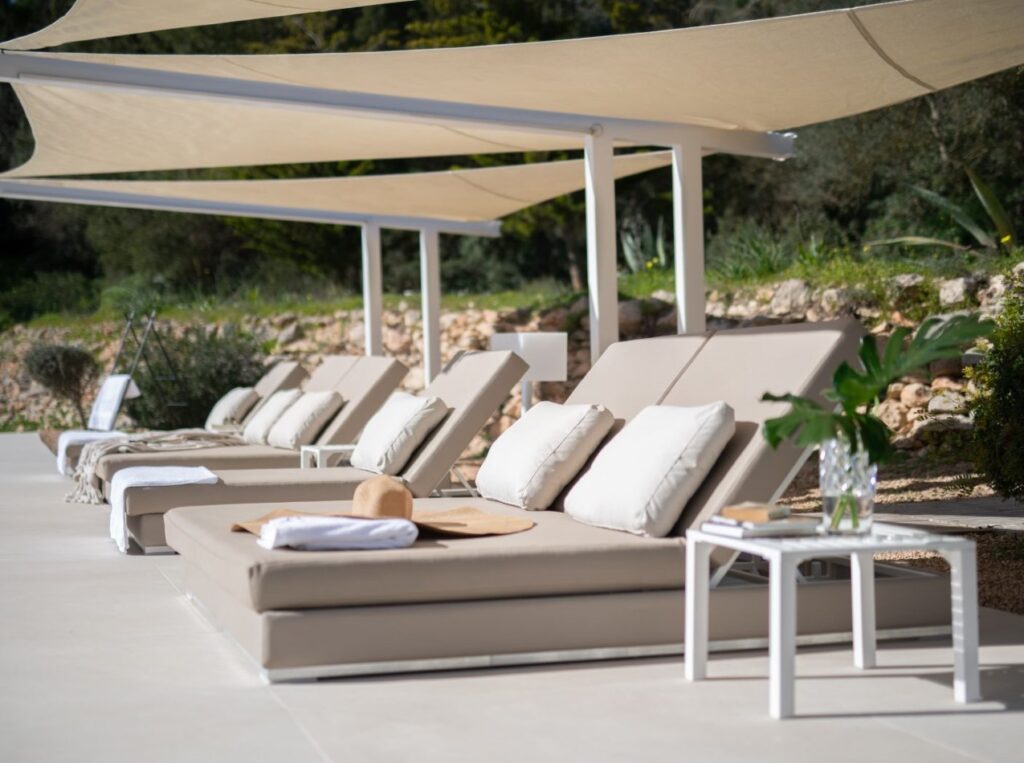 Lounge chairs with cushions are arranged under modern triangular shade sails in a sunlit outdoor space, embodying the essence of luxury travel. A small white table holds a vase with leaves, a sun hat, and a magazine. Greenery and rocks form the tranquil backdrop. David Pearson Travel