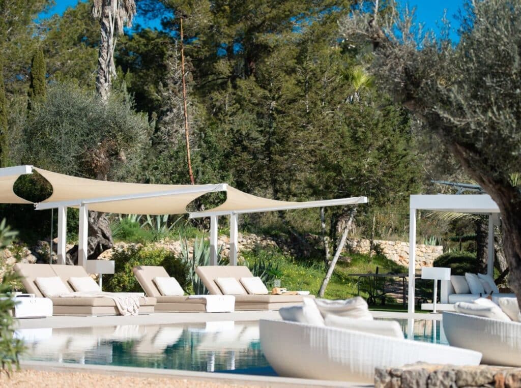 A serene outdoor pool area, perfect for luxury travel, is surrounded by lush greenery. Several lounge chairs with cushions are lined up under sunshades alongside a bright pool. White woven furniture graces the foreground, under a clear blue sky. David Pearson Travel