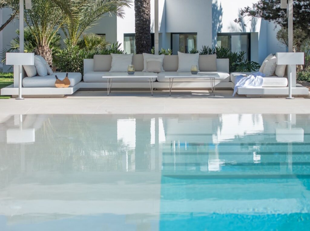 Experience the essence of luxury travel in a poolside oasis, where a modern outdoor sofa with beige cushions invites relaxation. Soft candlelight flickers on a nearby table, while a light blanket drapes over the side. A sleeping cat rests gracefully amid lush greenery and trees. David Pearson Travel