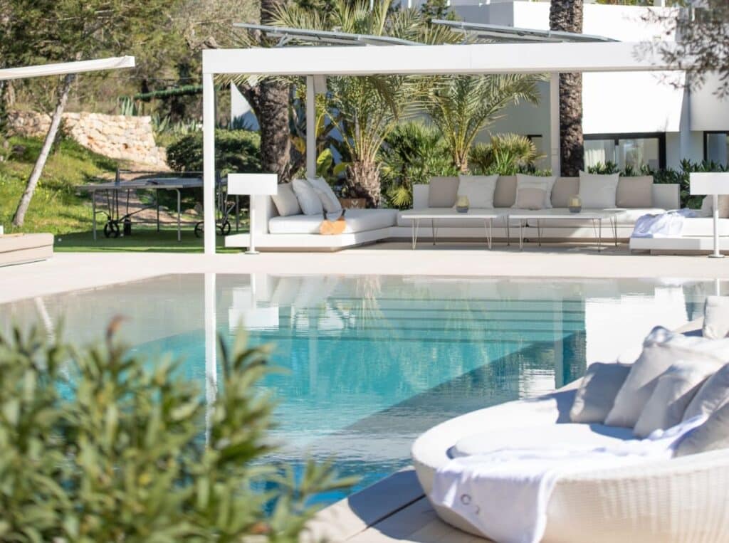 The luxurious outdoor pool area is perfect for luxury travel enthusiasts, featuring clear blue water, white cushioned lounge chairs, and a modern pergola. Palm trees and lush greenery create a serene, tropical atmosphere with a glimpse of a white building in the background. David Pearson Travel