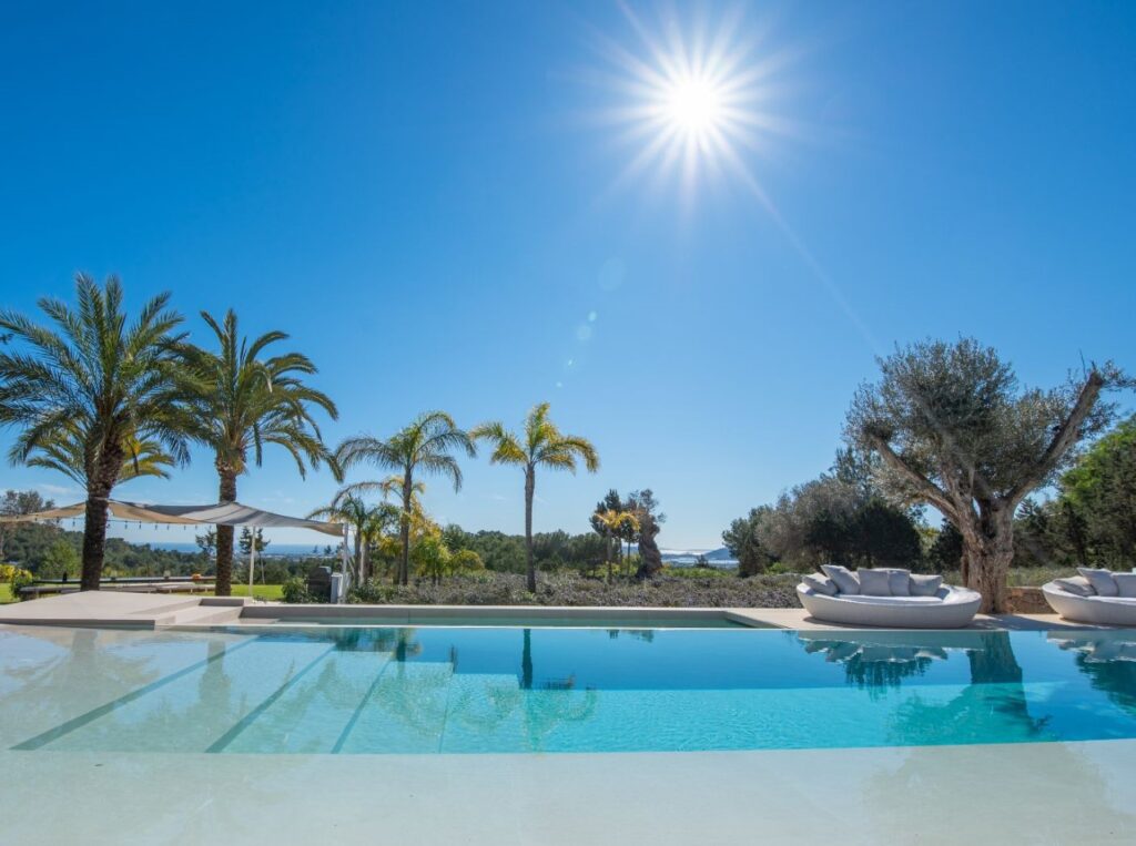 Experience luxury travel at its finest with a luxurious outdoor pool featuring clear blue water, surrounded by palm trees and lounge chairs. The sun shines brightly in a clear sky, casting reflections on the water, while scenic landscape views provide the perfect backdrop for relaxation. David Pearson Travel